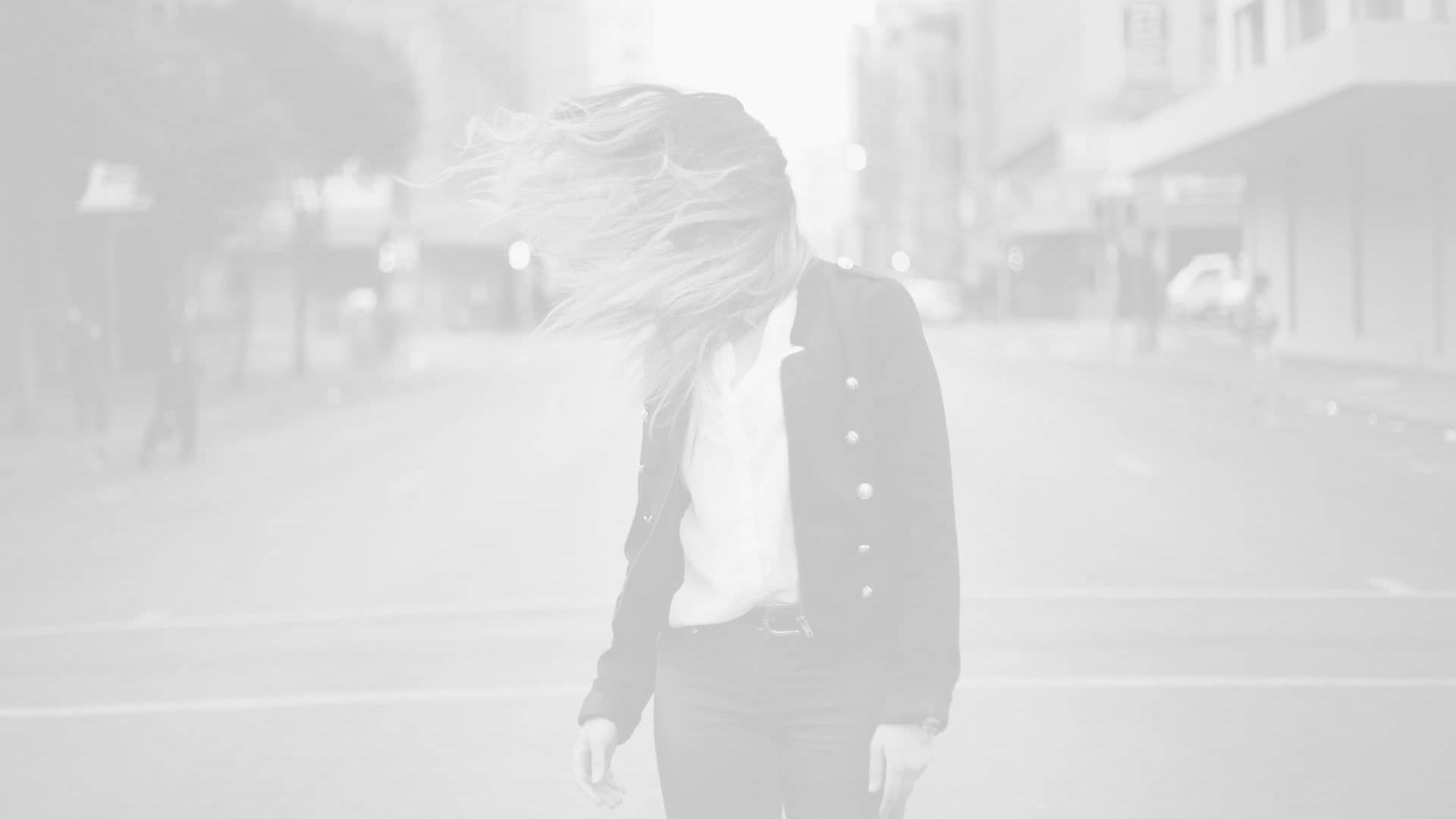 Person standing in the middle of a city street with their hair flipping forward, obscuring their face; buildings and cars are visible in the background.