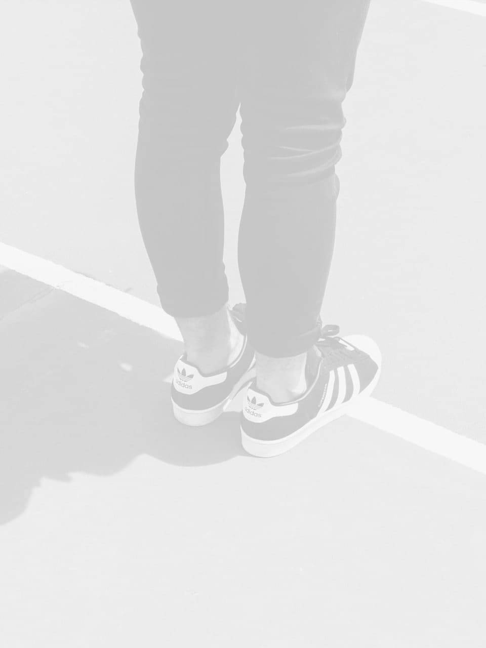 Person in black pants and Adidas sneakers standing on a track with visible white lines; their lower legs and shoes are shown with a shadow on the ground.