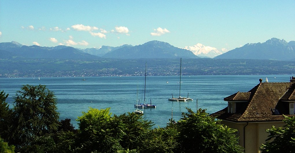 Blick auf einen ruhigen See mit Segelbooten, umrahmt von Bäumen und Dächern im Vordergrund und entfernten Bergen unter einem klaren blauen Himmel im Hintergrund.