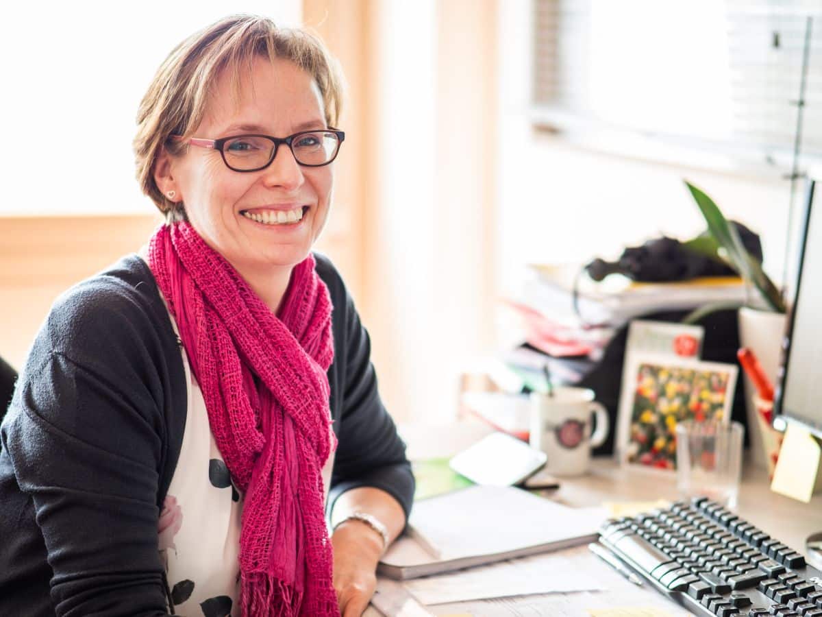 Une femme avec des lunettes et un foulard rose sourit tout en étant assise à un bureau avec des papiers, un clavier et des fournitures de bureau.