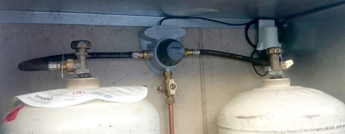 Zwei weiße Propangasflaschen, die mit schwarzen Schläuchen an einen Regler angeschlossen und an einer beigen Wand unter einem Metallregal installiert sind.