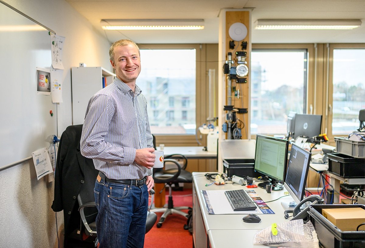 Ein Mann steht lächelnd in einem modernen Büro und hält eine Tasse neben einem Schreibtisch mit Computern und elektronischen Geräten.