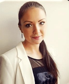 Eine Frau mit langen braunen Haaren in einem Pferdeschwanz, die einen weißen Blazer und ein schwarzes Oberteil trägt, steht vor einem schlichten hellen Hintergrund, blickt in die Kamera und lächelt leicht.