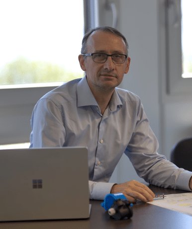 Un homme à lunettes est assis à un bureau avec un ordinateur portable, des documents et un petit appareil bleu, regardant la caméra dans un bureau lumineux.